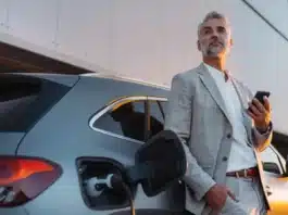 Businessman holding smartphone while charging car at electric vehicle charging station, closeup.